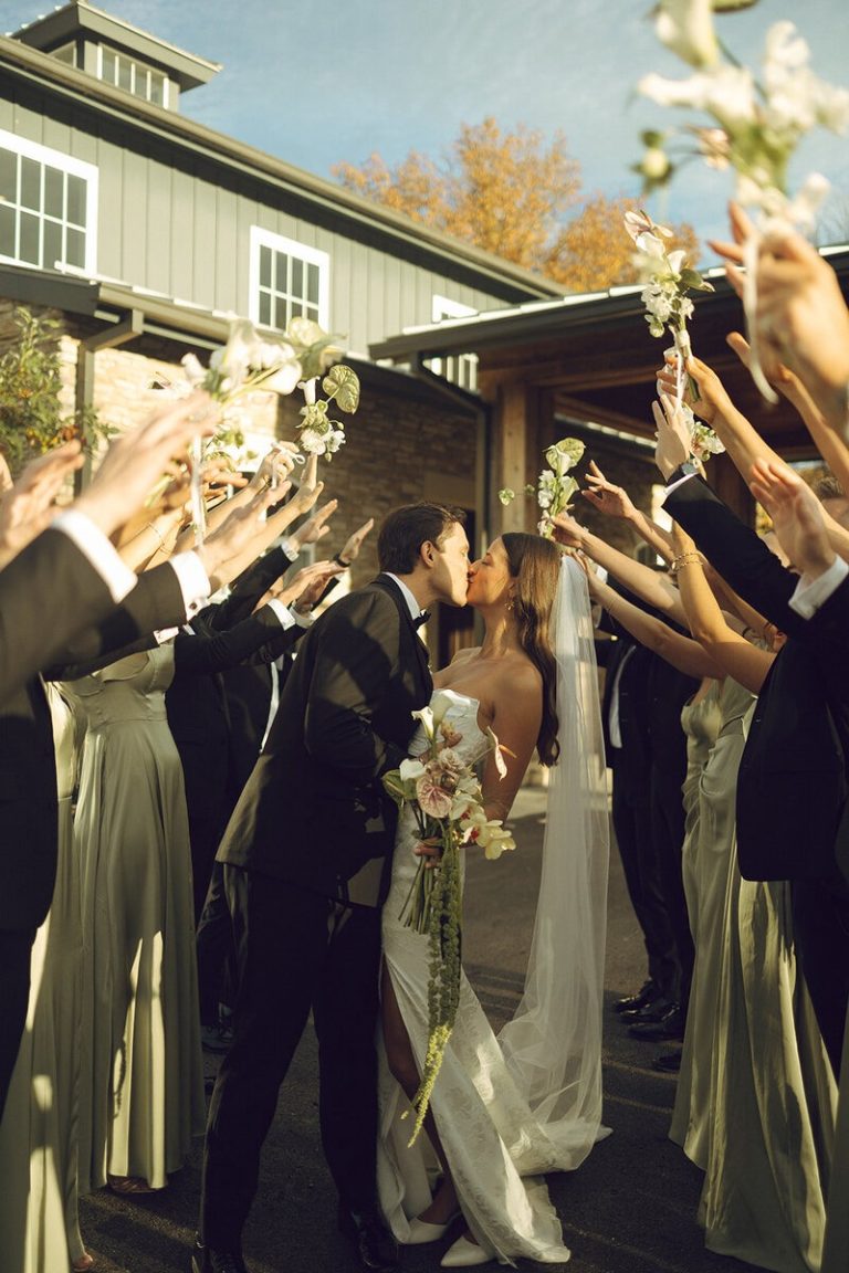 Bride and groom kiss beneath raised white flowers held by their wedding party outside a Columbus wedding venue.