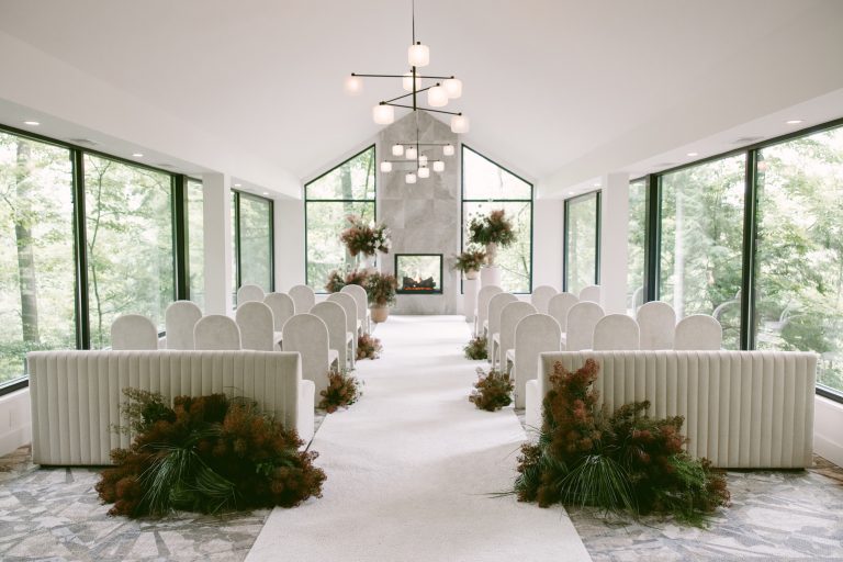 Earthy floral arrangements by Fiori Florals line a white ceremony aisle and frame a stone fireplace in a bright woodland setting.