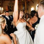 Bride in a strapless white wedding gown dances with guests during a lively reception at a rustic Columbus wedding venue with exposed wooden beams and warm lighting. Friends and family surround the dance floor, creating an energetic celebration with a packed crowd.