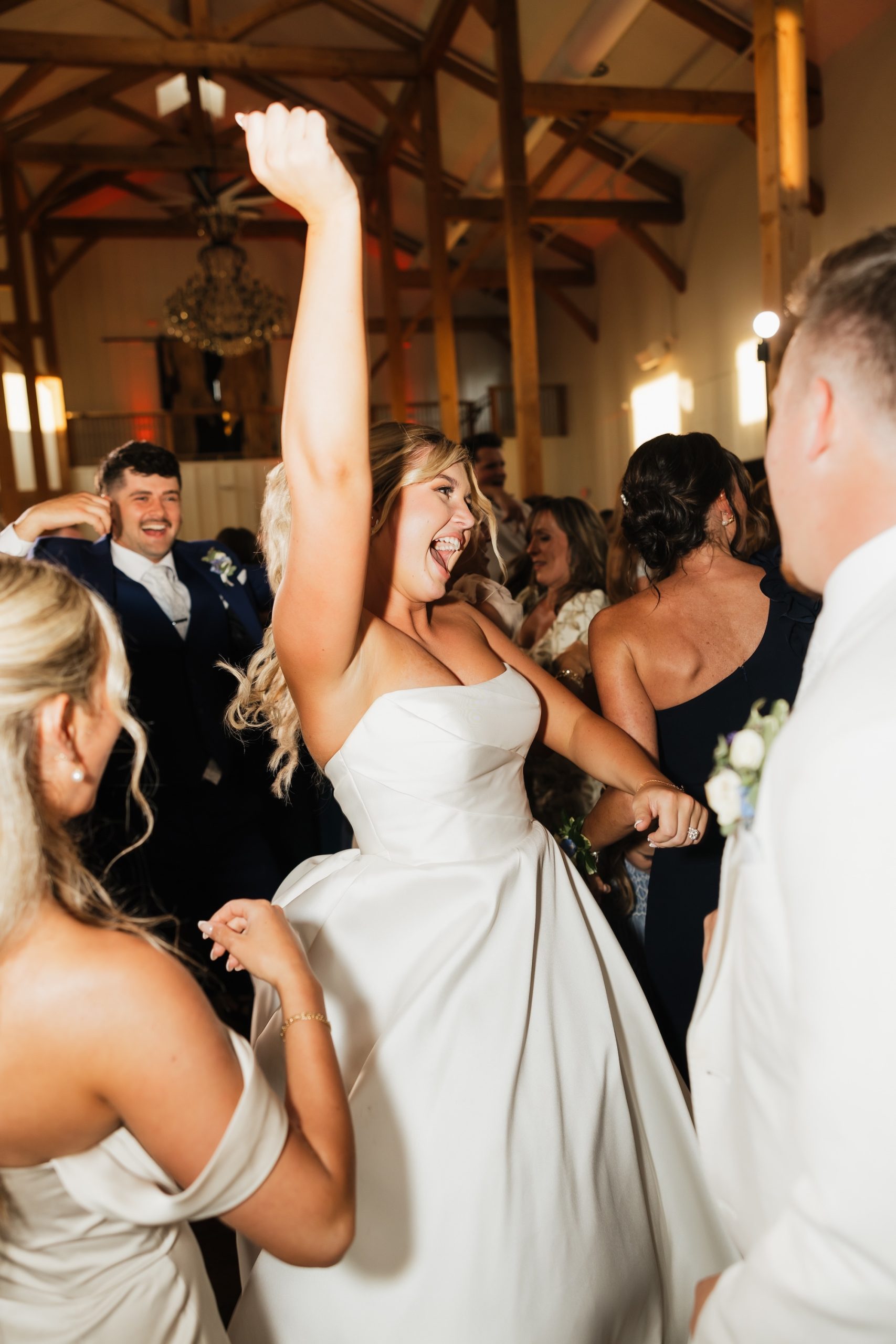 Bride in a strapless white wedding gown dances with guests during a lively reception at a rustic Columbus wedding venue with exposed wooden beams and warm lighting. Friends and family surround the dance floor, creating an energetic celebration with a packed crowd.