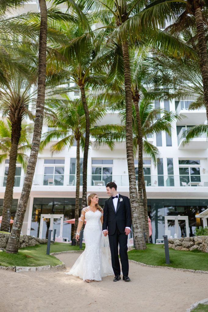 Bride and groom holding hands under palm trees at modern waterfront venue