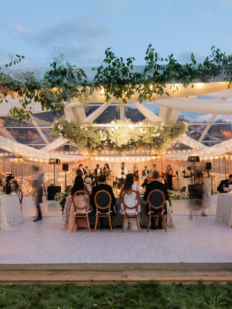 Elegant evening wedding reception under illuminated clear-top tent with hanging greenery, string lights, and guests dining at formal tables