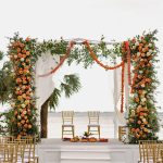 Waterfront wedding ceremony setup with white draped canopy adorned with orange and yellow floral garlands, gold chiavari chairs, and white platform