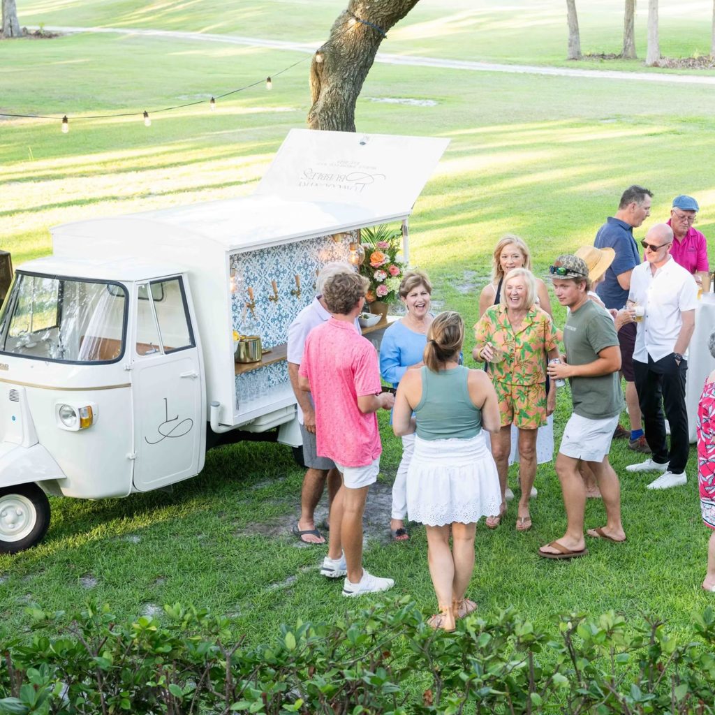 Wedding guests gathering around white vintage mobile bar trailer on outdoor lawn
