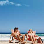 Couple relaxing in striped beach chairs on sandy shore, holding hands in sunglasses