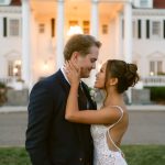 Bride and groom share an intimate moment at sunset in front of an elegant illuminated white mansion venue