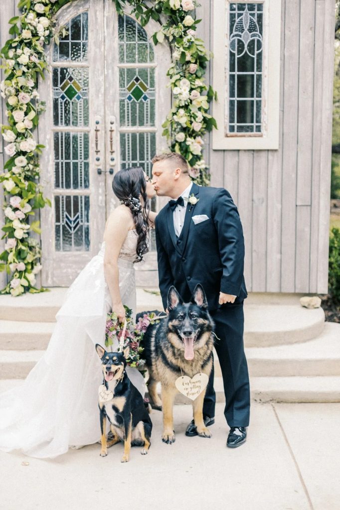 Bride and groom kissing with two dogs wearing floral collars in front of rustic chapel entrance