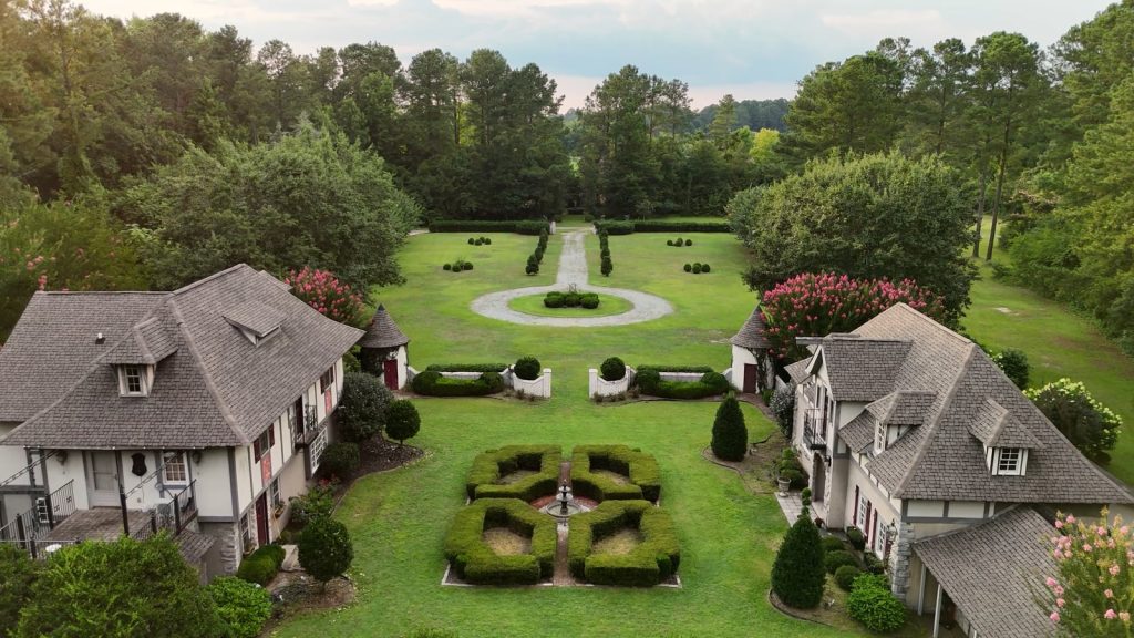 Aerial view of formal European garden estate with symmetrical lawns, circular fountain, topiary, and twin villas flanking manicured grounds
