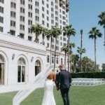 Bride and groom walking hand-in-hand on manicured lawn outside elegant white hotel with palm trees and ceremony seating