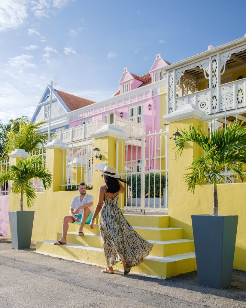 Woman in flowing dress walking up yellow stairs at tropical resort with pastel-colored buildings