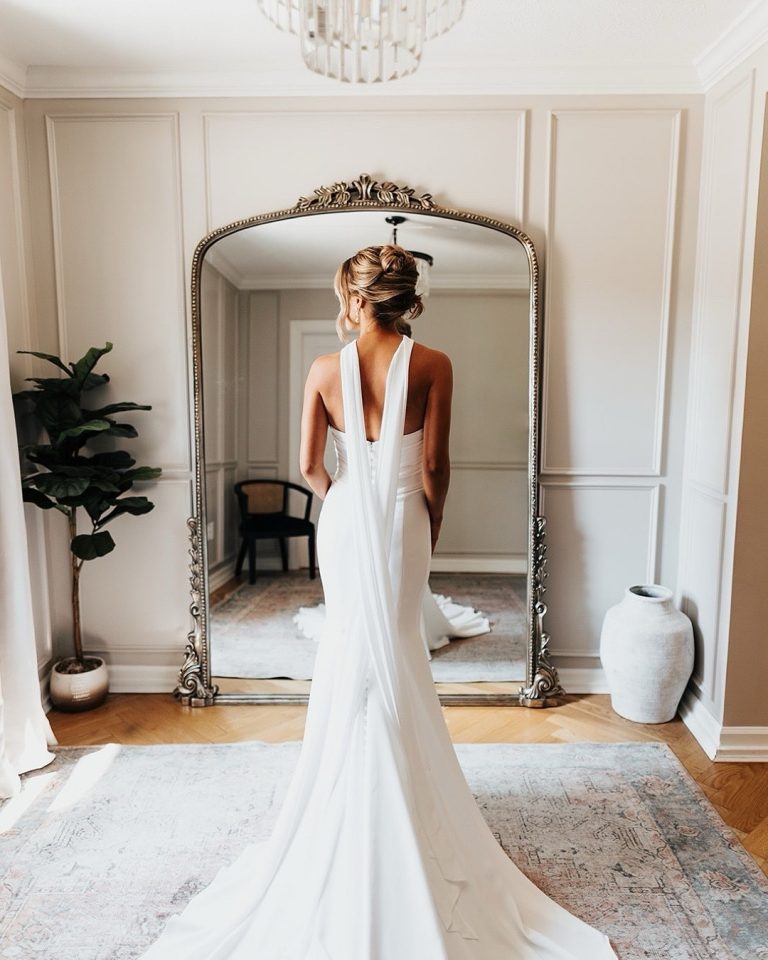 Bride in sleek white halter gown with low back standing before ornate gold-framed mirror