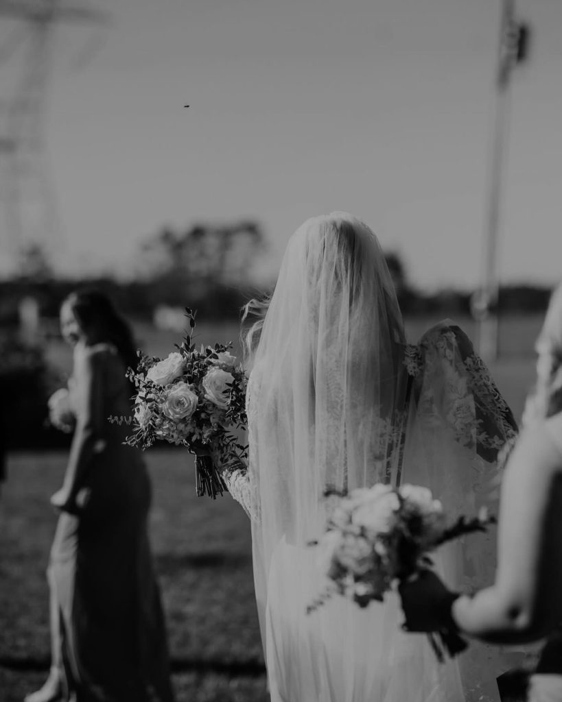 Bride in veil holding rose bouquet walking down outdoor ceremony aisle with bridesmaids