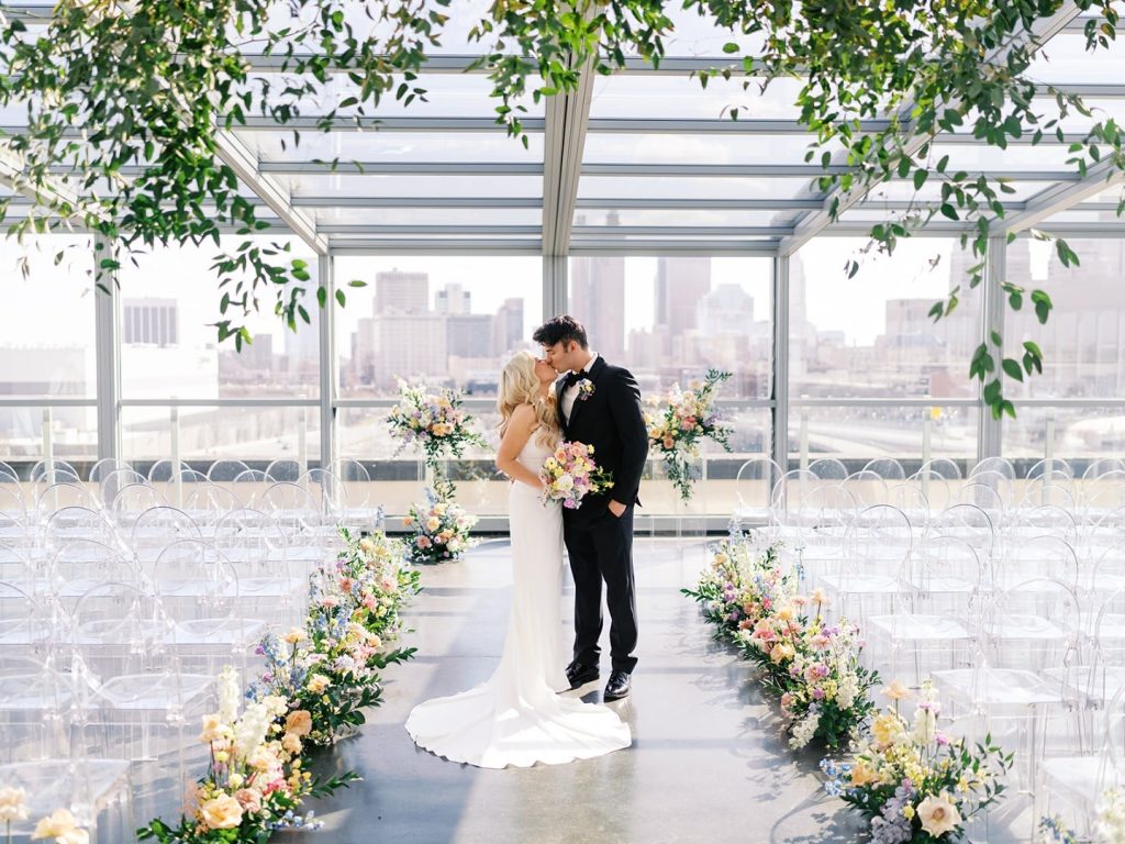 Couple embraces in modern glass-walled ceremony space decorated with greenery and romantic lighting