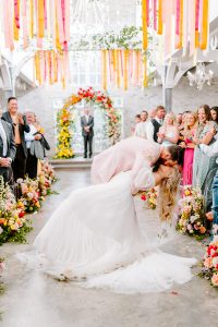 Newlyweds kiss under colorful ribbon streamers and floral arch at vibrant ceremony