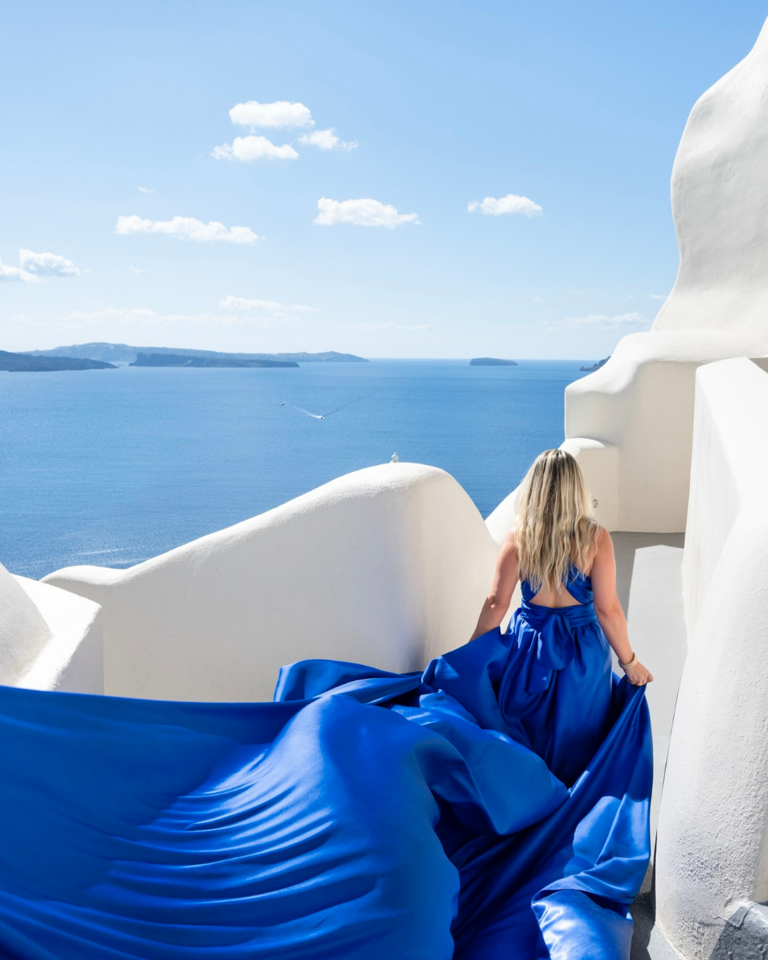 Woman in flowing royal blue dress on white Santorini terrace overlooking Aegean Sea, exemplifying destination wedding elegance