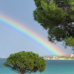 Rainbow arcing over turquoise Mediterranean waters with beach umbrellas and pine trees, showcasing a luxury honeymoon destination