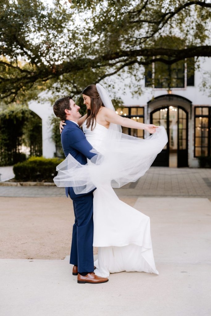 Groom in navy suit lifts bride in flowing white dress and veil beneath sprawling oak tree at Houston venue
