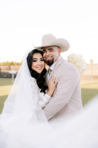 Smiling bride and groom in cowboy hat embrace outdoors holding white bridal bouquet