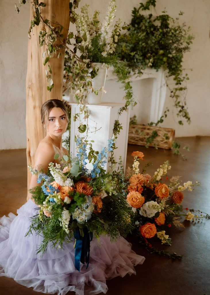 Bride in flowing white gown holds vibrant bouquet with orange, blue, and white blooms against natural greenery backdrop