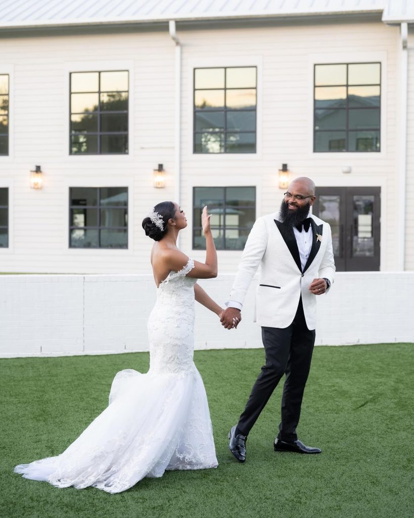 Bride and groom holding hands and dancing on lawn outside modern white venue with black-framed windows