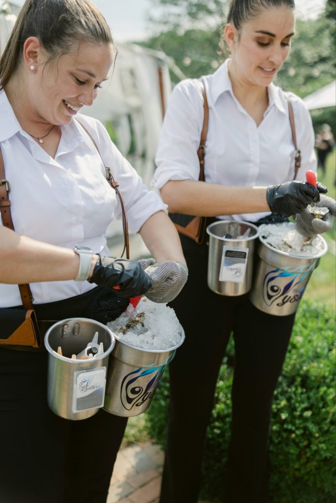 Blue Ember Oysters team members shucking fresh oysters during an outdoor wedding cocktail hour in Columbus, Ohio. The interactive catering experience features roaming oyster servers preparing and serving oysters on ice for wedding guests.