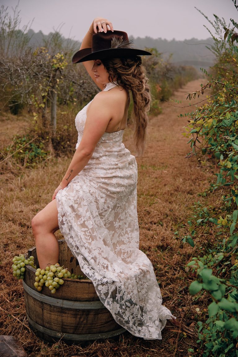 Bride in lace gown and wide-brim hat sitting on wine barrel among vineyard rows