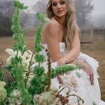 Bride in strapless white gown surrounded by lush floral arrangements with vineyard landscape in background