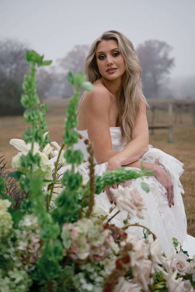 Bride in strapless white gown surrounded by lush floral arrangements with vineyard landscape in background
