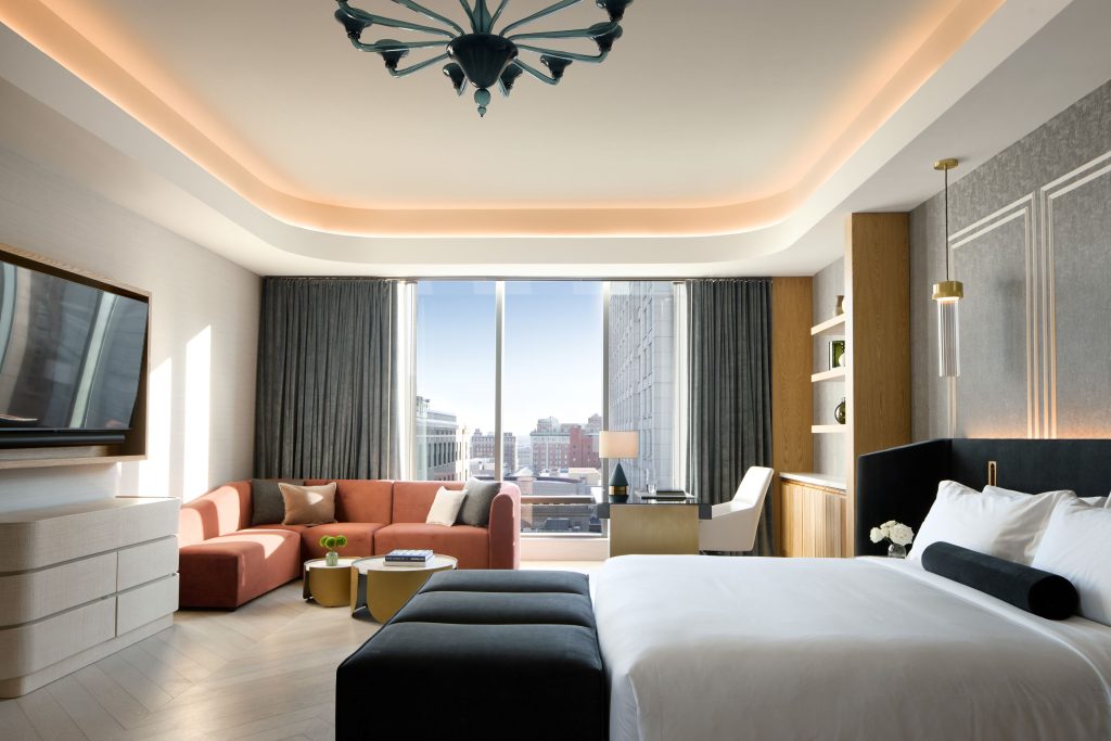 Spacious hotel suite featuring king bed, coral sectional sofa, butterfly chandelier, and floor-to-ceiling windows overlooking downtown Indianapolis