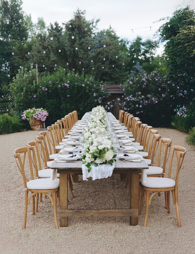 Long outdoor garden reception table with wooden farm table, bentwood chairs, white floral runner, and string lights overhead