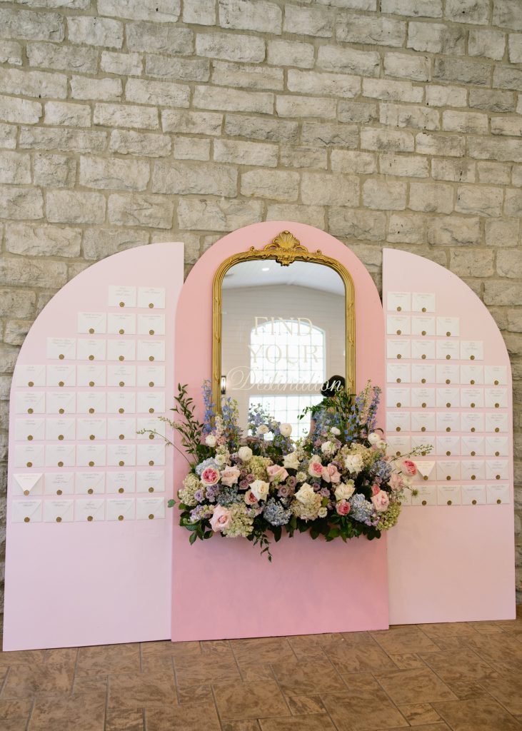 Pink arched seating chart backdrop adorned with mirror and romantic floral arrangement