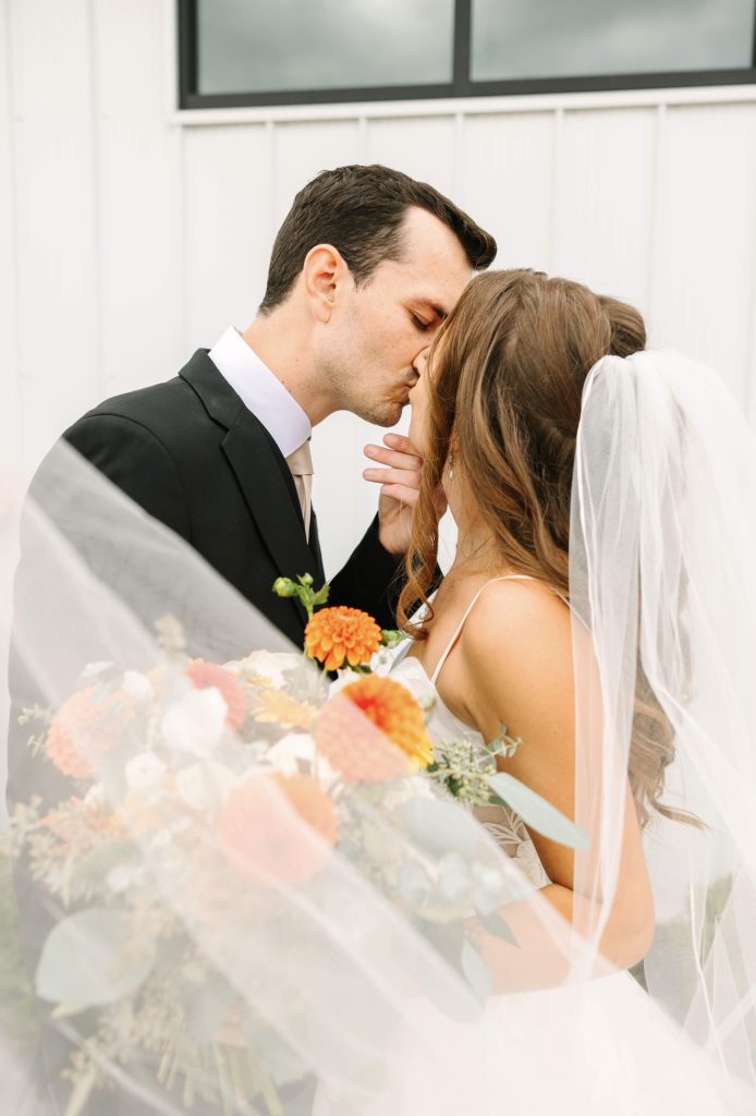 Newlyweds kissing wrapped in flowing veil with colorful bouquet between them