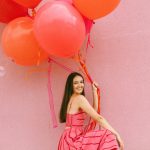 Woman in coral pink gown holds cluster of orange and pink balloons against matching pink wall