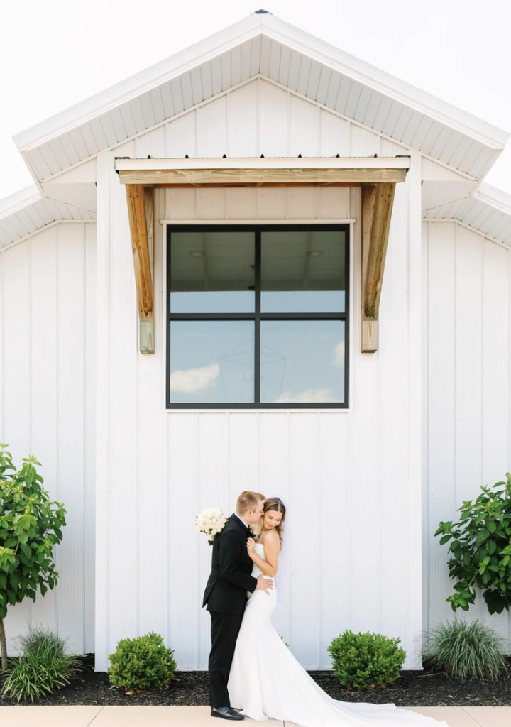 Bride and groom embrace in front of white modern barn venue with peaked roof and black-framed windows