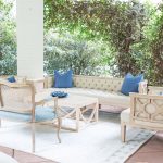 Outdoor wedding lounge area featuring cream tufted sofa, whitewashed cane-back chairs, and blue accent pillows surrounded by lush greenery