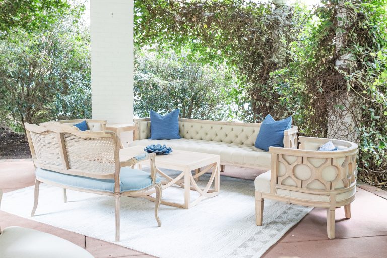 Outdoor wedding lounge area featuring cream tufted sofa, whitewashed cane-back chairs, and blue accent pillows surrounded by lush greenery
