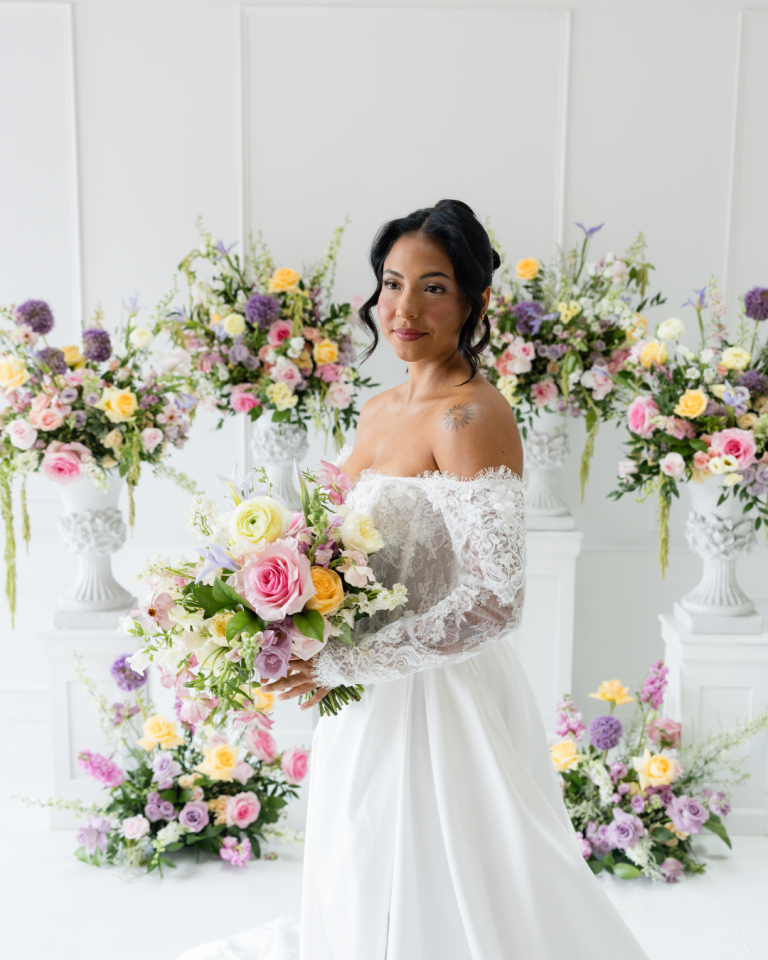 Bride in off-shoulder lace wedding gown holding colorful bouquet with pink, yellow, and purple blooms surrounded by matching floral arrangements