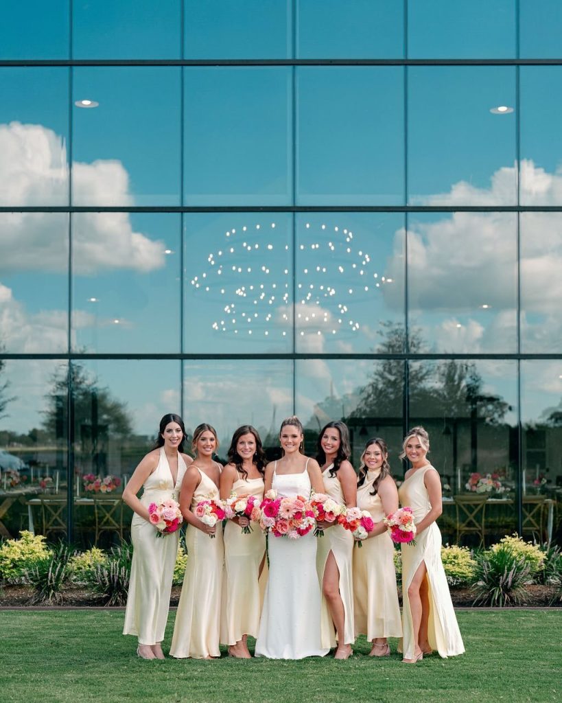 Bride and bridesmaids in champagne gowns hold pink bouquets outside modern glass venue with white balloons