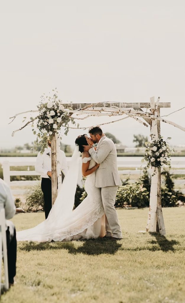 Newlyweds embracing beneath floral wedding arch with white fence and open pasture in background at Rockin' S Ranch