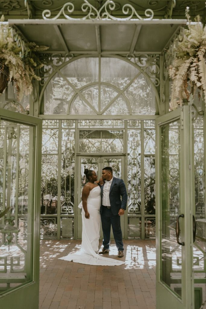 Bride and groom embracing in elegant glass conservatory with ornate white framework and natural light