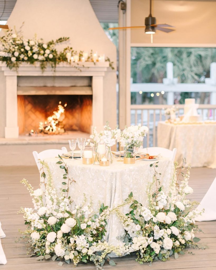 Elegant reception table with white lace linens, white floral garland, and candlelight beside fireplace