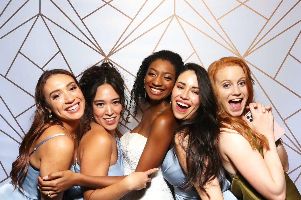 Bride and bridesmaids in blue dresses laughing together at wedding photo booth with geometric gold backdrop