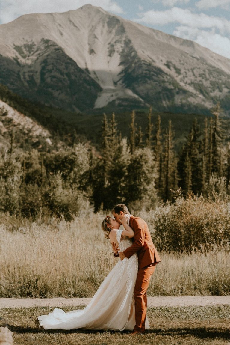 Bride and groom kissing in golden meadow with dramatic Rocky Mountain peak and pine forest backdrop