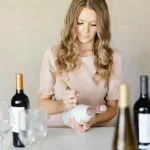 Woman in blush dress personalizing wine bottle with gold pen at experiential wedding wine tasting station