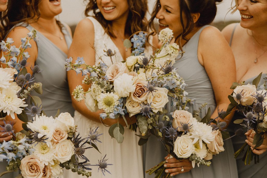 Bridesmaids in sage green gowns holding bouquets with cream blooms and blue accents