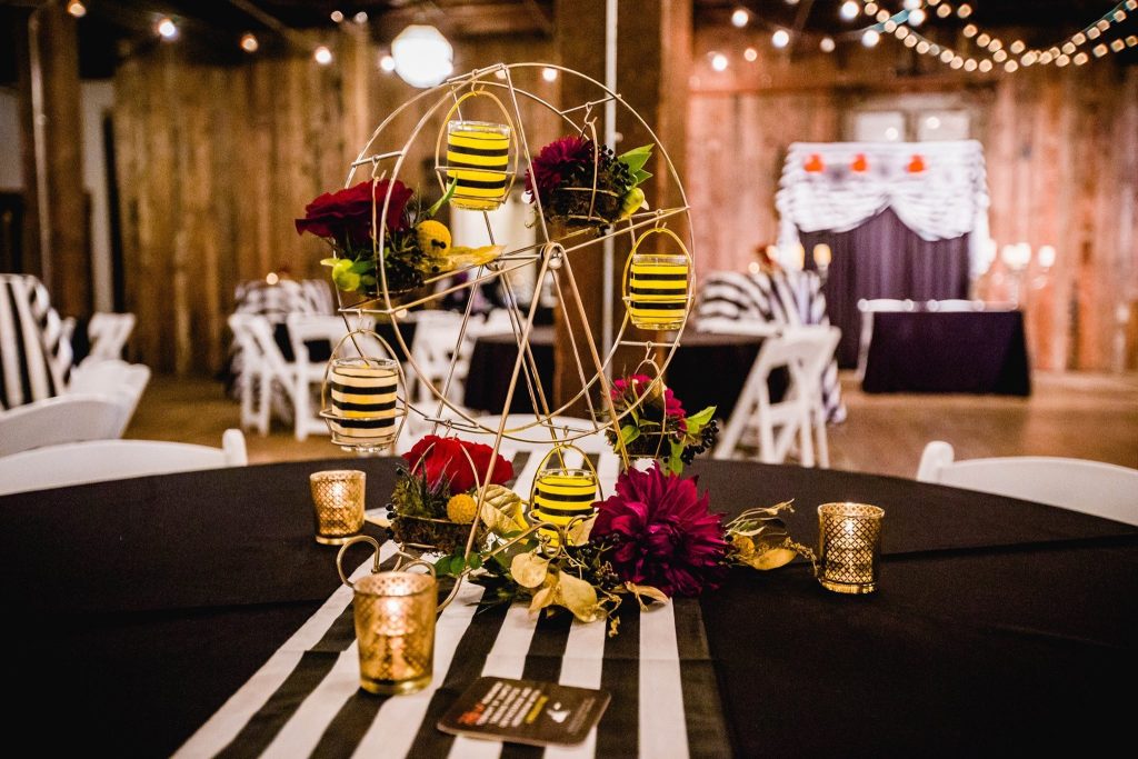 Whimsical Ferris wheel centerpiece with bee-themed decorations, burgundy flowers, and gold candles on reception table in rustic barn venue