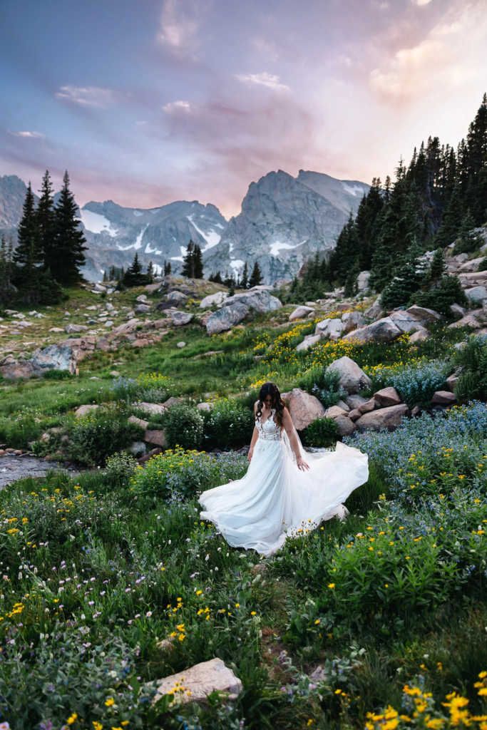 Bride twirling in flowing white gown among wildflowers in alpine meadow with mountain peaks at sunset
