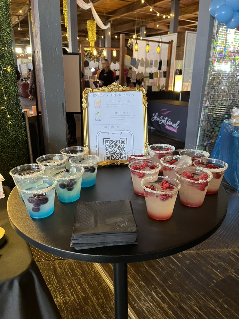 Mobile bar display featuring blue and red cocktails with gold-framed QR code menu sign at wedding event