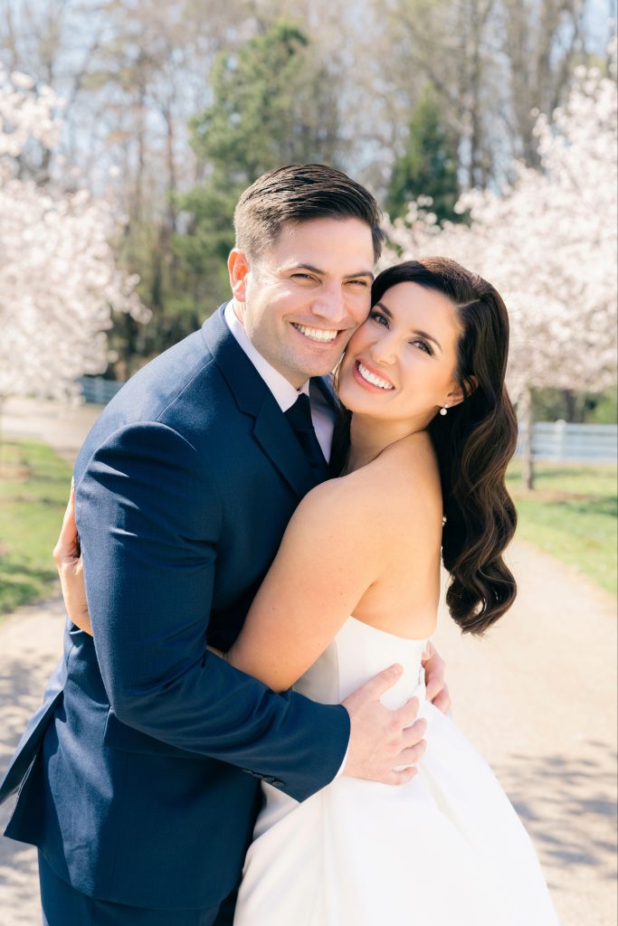 Smiling bride and groom embracing in navy suit and white wedding dress among spring cherry blossoms