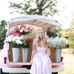 Woman in pink gingham dress sitting at vintage pink flower truck displaying fresh blooms in galvanized buckets